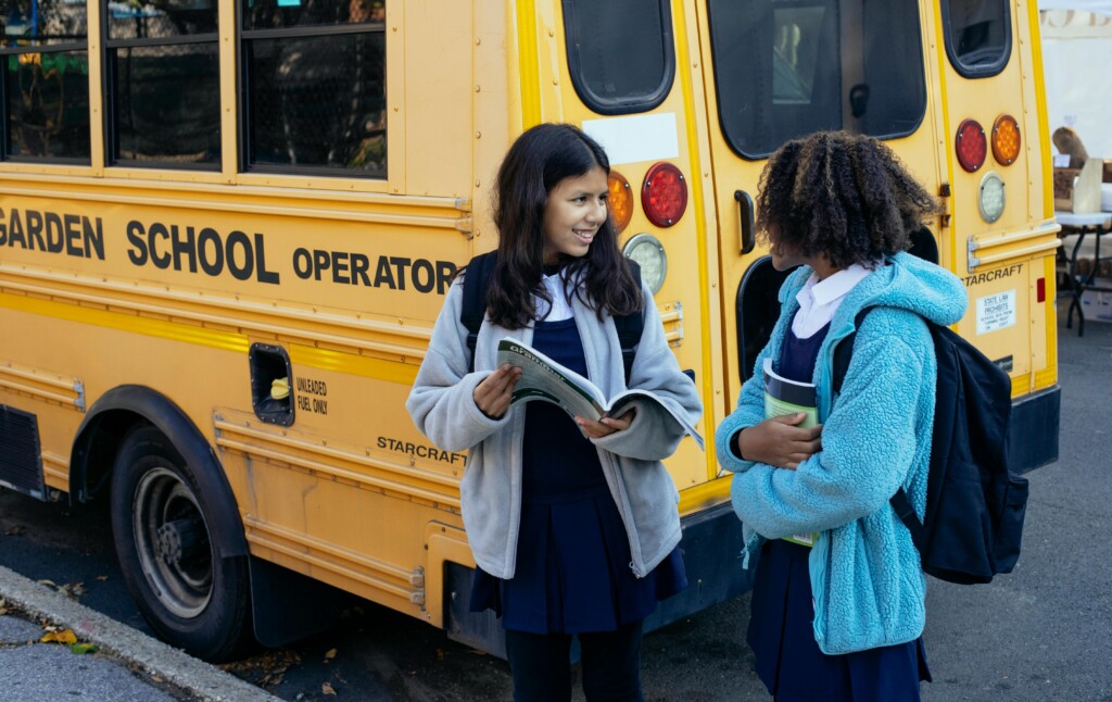 Two students standing by a yellow school bus discussing a book.