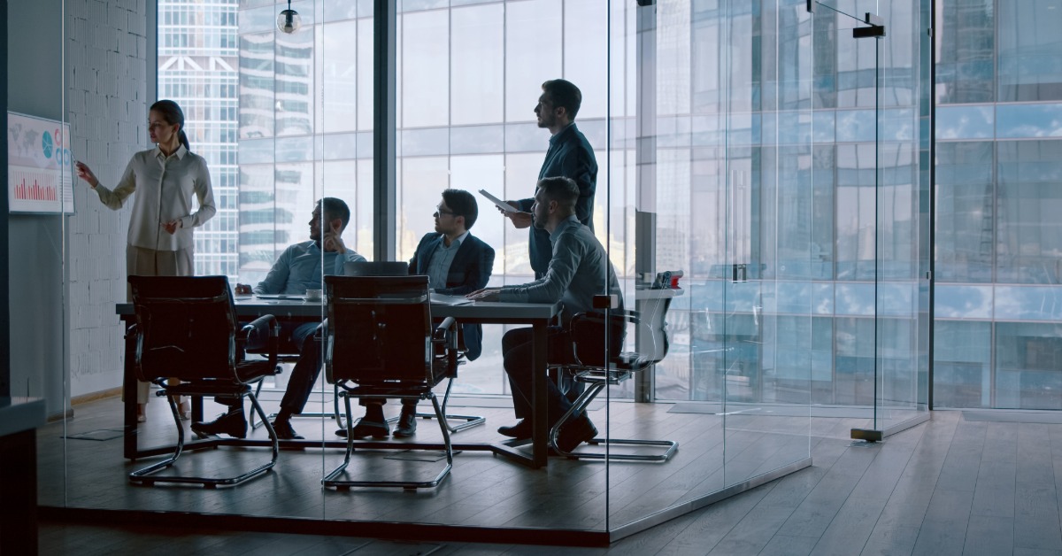 Four coworkers stand by tall windows in an office building and review a clipboard held between them.