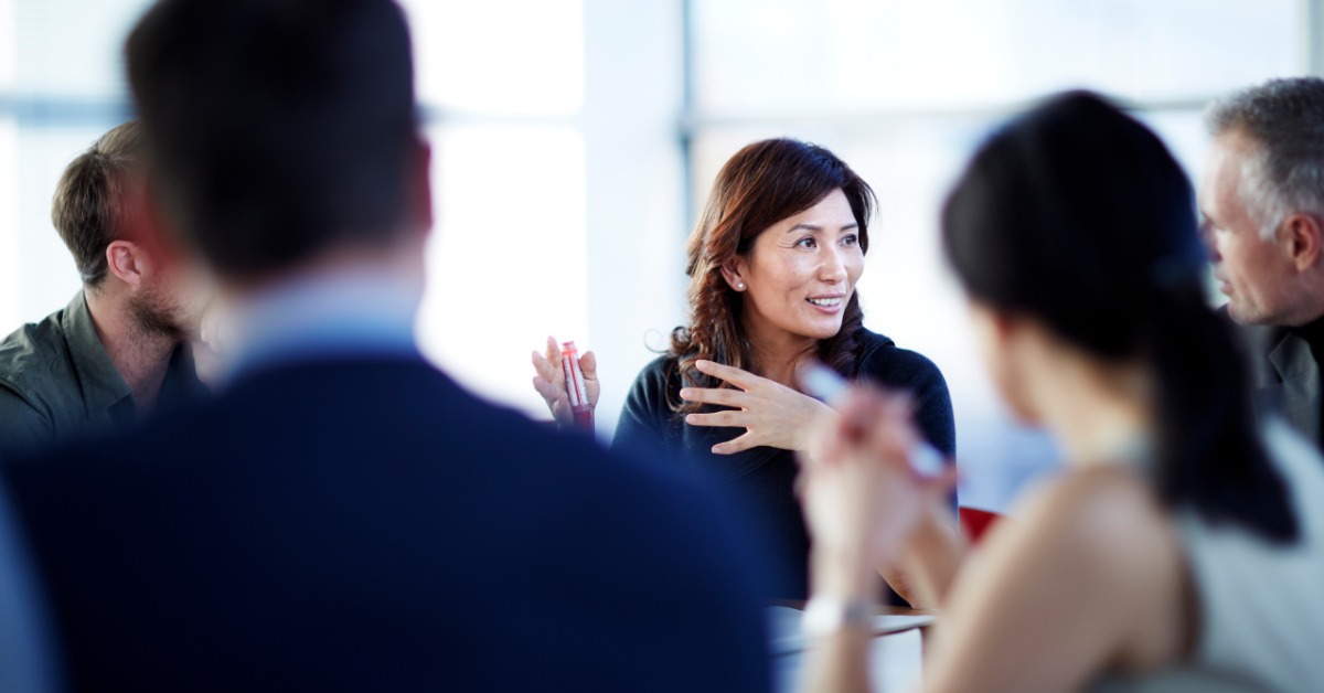 Five coworkers engaged in discussion around a conference table in a bright office. A smiling, gesturing woman is in focus.