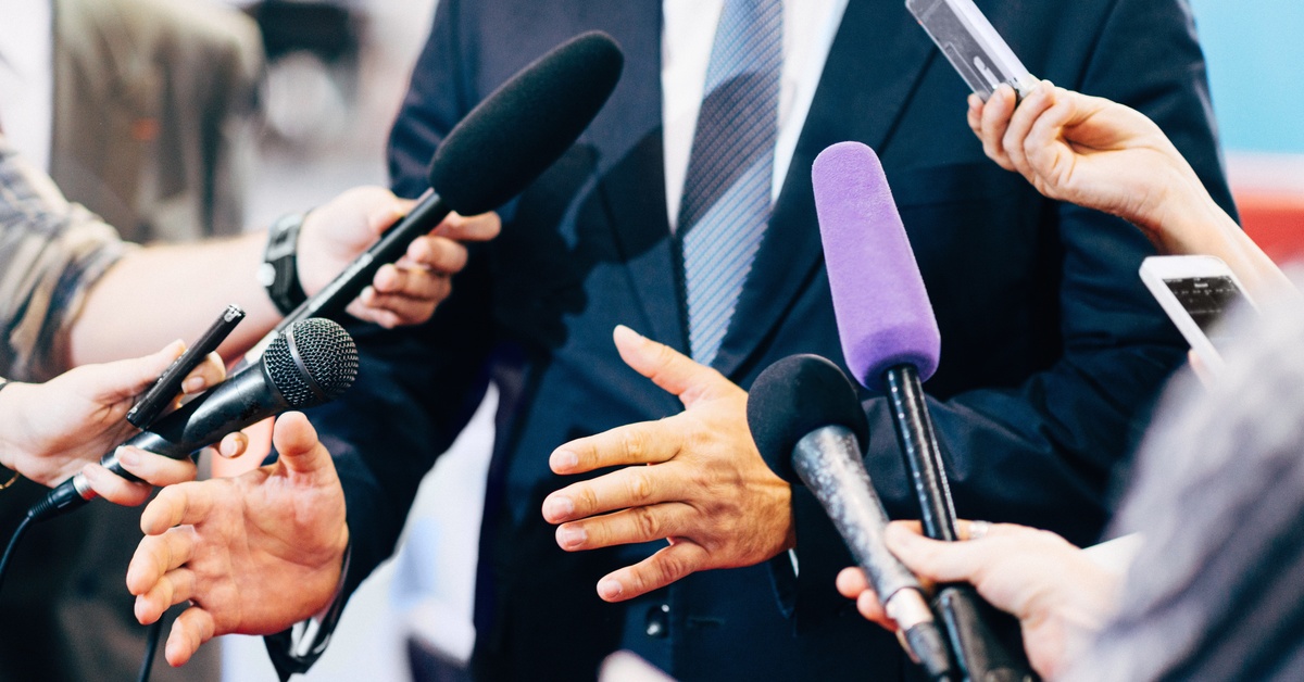 A man in a suit gestures with his hands as he speaks to reporters holding microphones and recording devices all around him.
