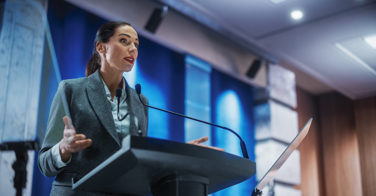 A woman in a gray blazer speaks at a podium with two microphones in a conference hall, gesturing with both hands.