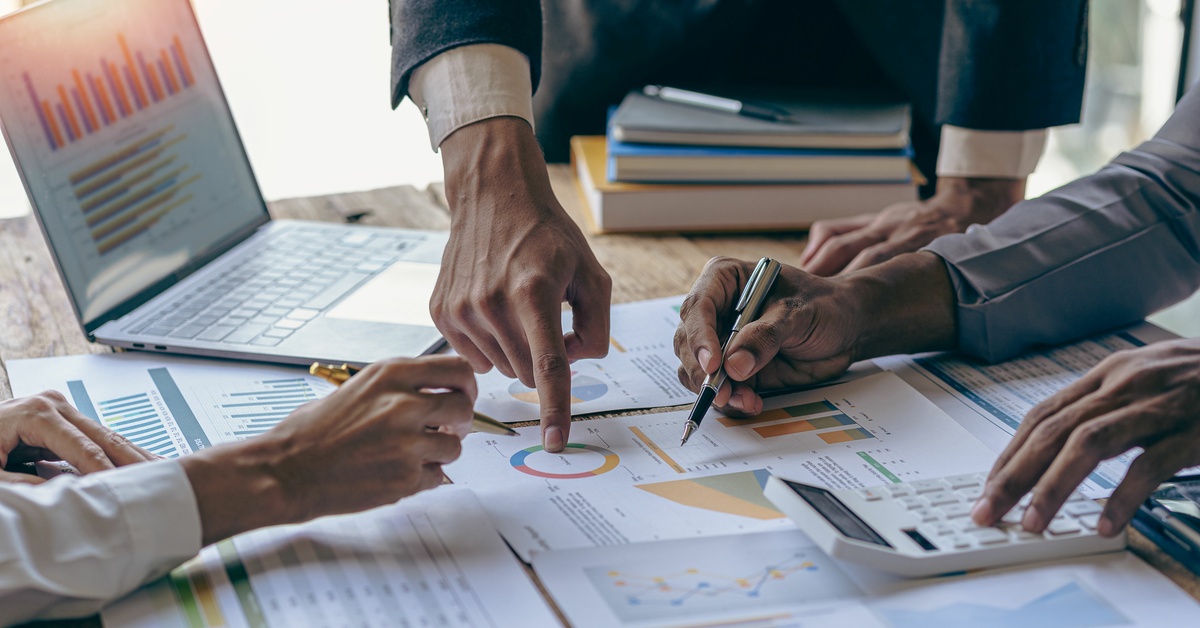 A close-up of hands pointing to printed charts and graphs on a desk beside a laptop, calculator, and books.
