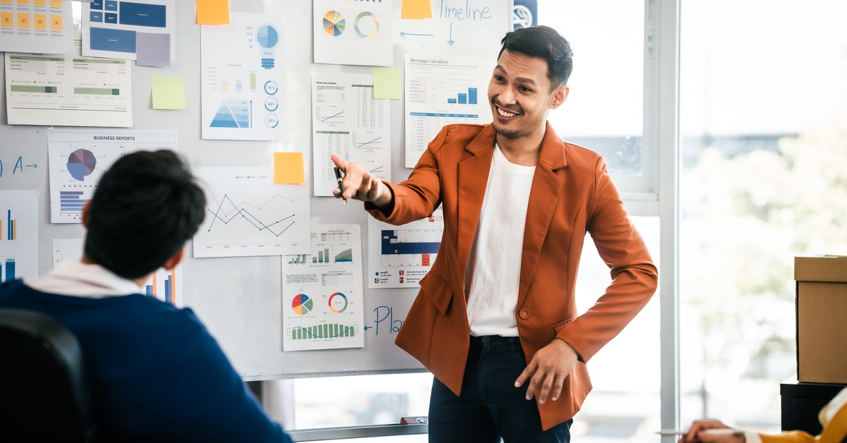 A smiling man in an orange blazer presents charts on a whiteboard to his coworkers in a sunlit meeting room.
