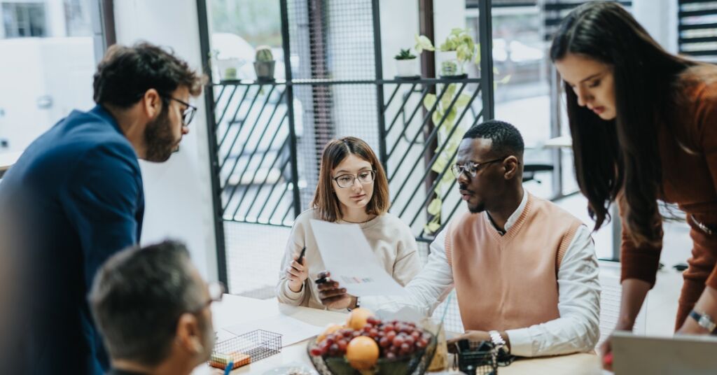 A diverse team gathers around a table in a bright office, reviewing notes during a collaborative meeting.