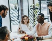A diverse team gathers around a table in a bright office, reviewing notes during a collaborative meeting.