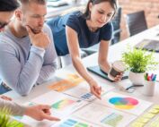 Four concentrated coworkers gather around a table and examine the printed charts and sticky notes organized on it.