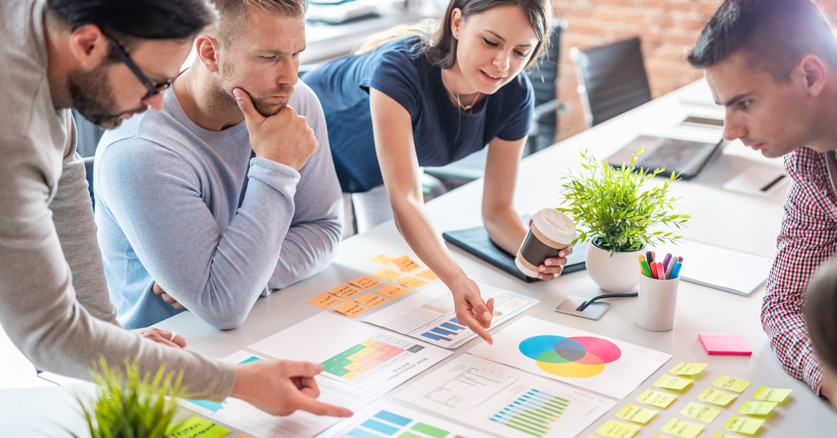 Four concentrated coworkers gather around a table and examine the printed charts and sticky notes organized on it.
