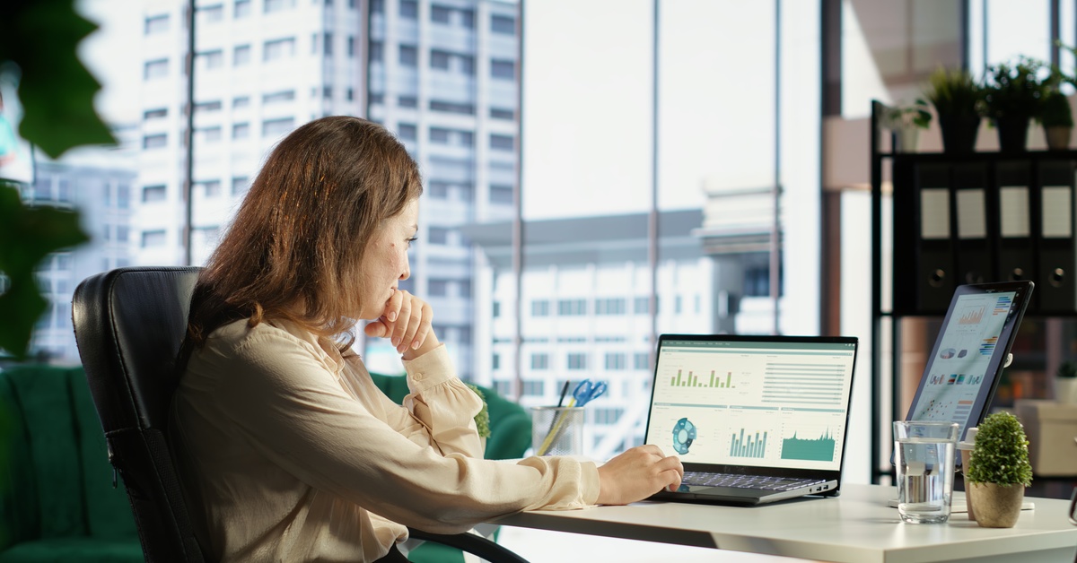 A concentrated woman in a beige blouse sits at her city office desk and looks at charts on a laptop.